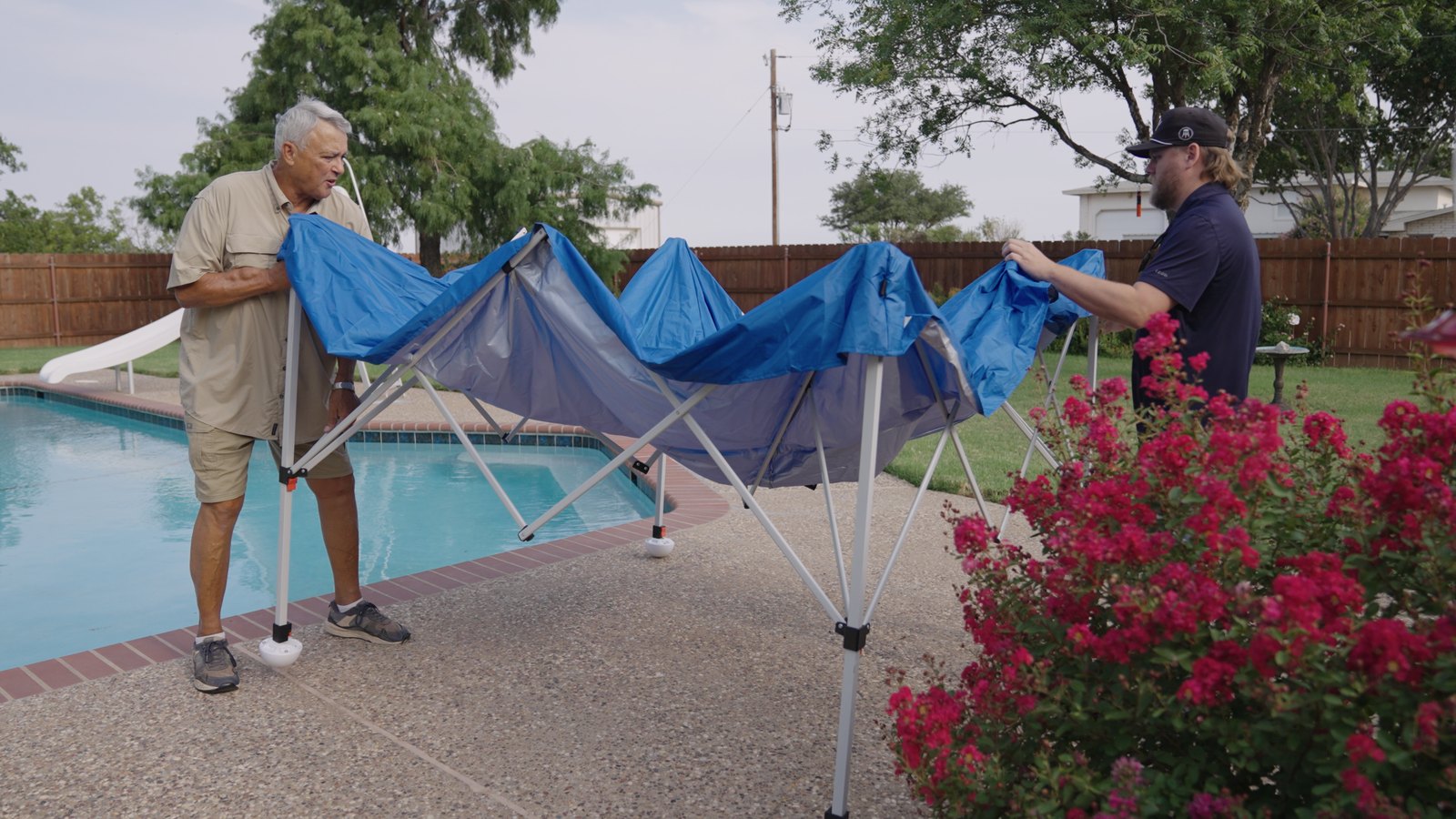 Two men setting up a 10×10 pop-up canopy as pool shade at the edge of a backyard swimming pool, Canopy Turtles bolted onto each leg.