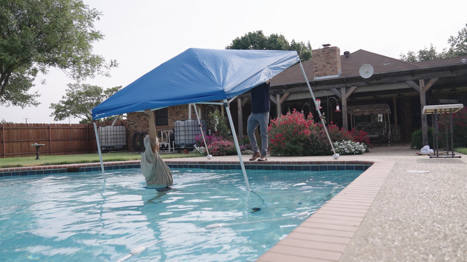 Pool shade in action — a 10×10 pop-up canopy standing directly inside an in-ground swimming pool with the legs protected by Canopy Turtles.